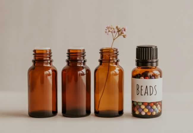 Repurposed essential oil bottles showing creative reuse options: clean amber glass bottles used as a mini bud vase with delicate dried flowers and as craft supply storage container filled with colorful beads, demonstrating practical second-life applications.